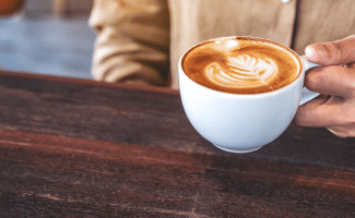 a person sits at a wooden table holding an artfully poured latte. we can only see their hand and the front of their shirt.