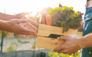 A man's hands recieve an apple crate full of fresh produce from another man wearing an apron.