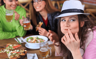 Woman with a hat looks just past the camera, a slight smile on her face, which is full of delicious food. Her friends laugh in the background.