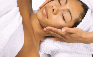 A woman lies down with her eyes closed. her body is wrapped in a towel, her head in another. she looks peaceful as two hands gently massage her neck.