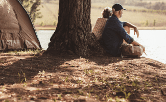 An older couple looks out on a lake as they sit next to an old pine tree. Their tent is in the background.