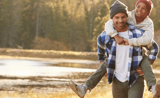 A man in a flanel shirt and beanie carries a boy on his back. They've been on a long hike and the sun is setting.