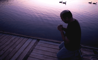 a mustachioed man prays in the early morning light on a dock. four ducks swim by in the background.