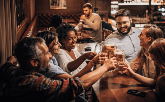 a group of diverse friends "cheers" with glasses of beer at a pub. a couple in the background might be on a date!
