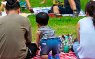 A family of three sits on a red and white checkered picnic blanket on the grass. There are other families in the background enjoying the beautiful day.