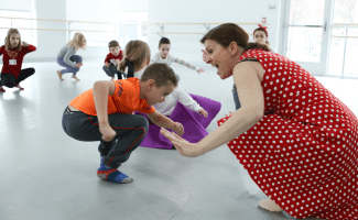 An enthusiastic teacher in a polkadotted red dress leads dance moves to a group of young elementary school aged children.