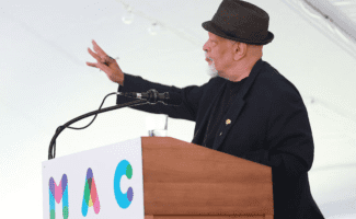 A man speaks from behind a podium. He wears a fedora and black suit and holds his glasses in his hand as he gestures.
