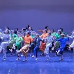 a stage full of kids dancing in different colored costumes under blue lighting