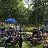 people lounging on grass with chairs, blankets, and umbrellas facing the concert