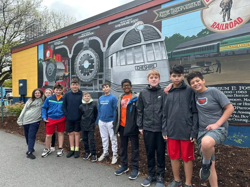 a group of middle schoolers smile in front of a mural of a train