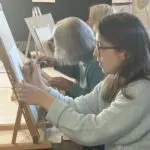 A group of artists works on easels in a classroom setting.