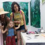 Two people hold the hands of a young child as they explore a colorful artist studio. tools and works in progress lay on the table next to them.