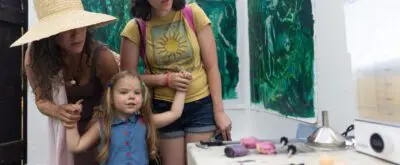 Two people hold the hands of a young child as they explore a colorful artist studio. tools and works in progress lay on the table next to them.