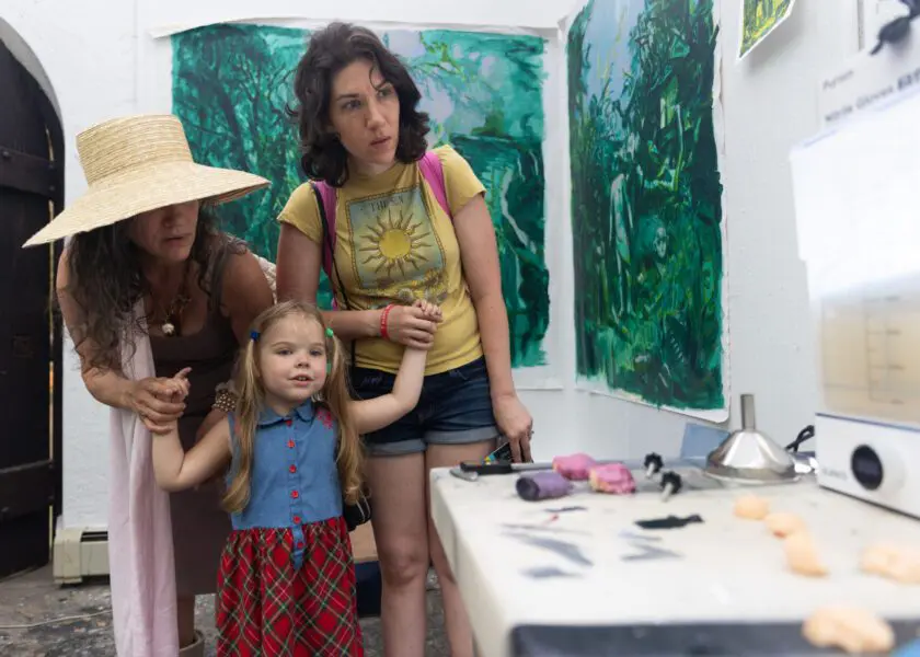 Two people hold the hands of a young child as they explore a colorful artist studio. tools and works in progress lay on the table next to them.