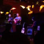 three musicians perform at the coffee house stage of Nova Arts, surrounded by art installation of glowing whales suspended from the ceiling