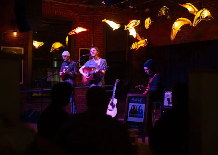 three musicians perform at the coffee house stage of Nova Arts, surrounded by art installation of glowing whales suspended from the ceiling