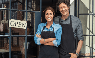 A couple stands proudly in front of an open sign. They both wear aprons and big smiles.